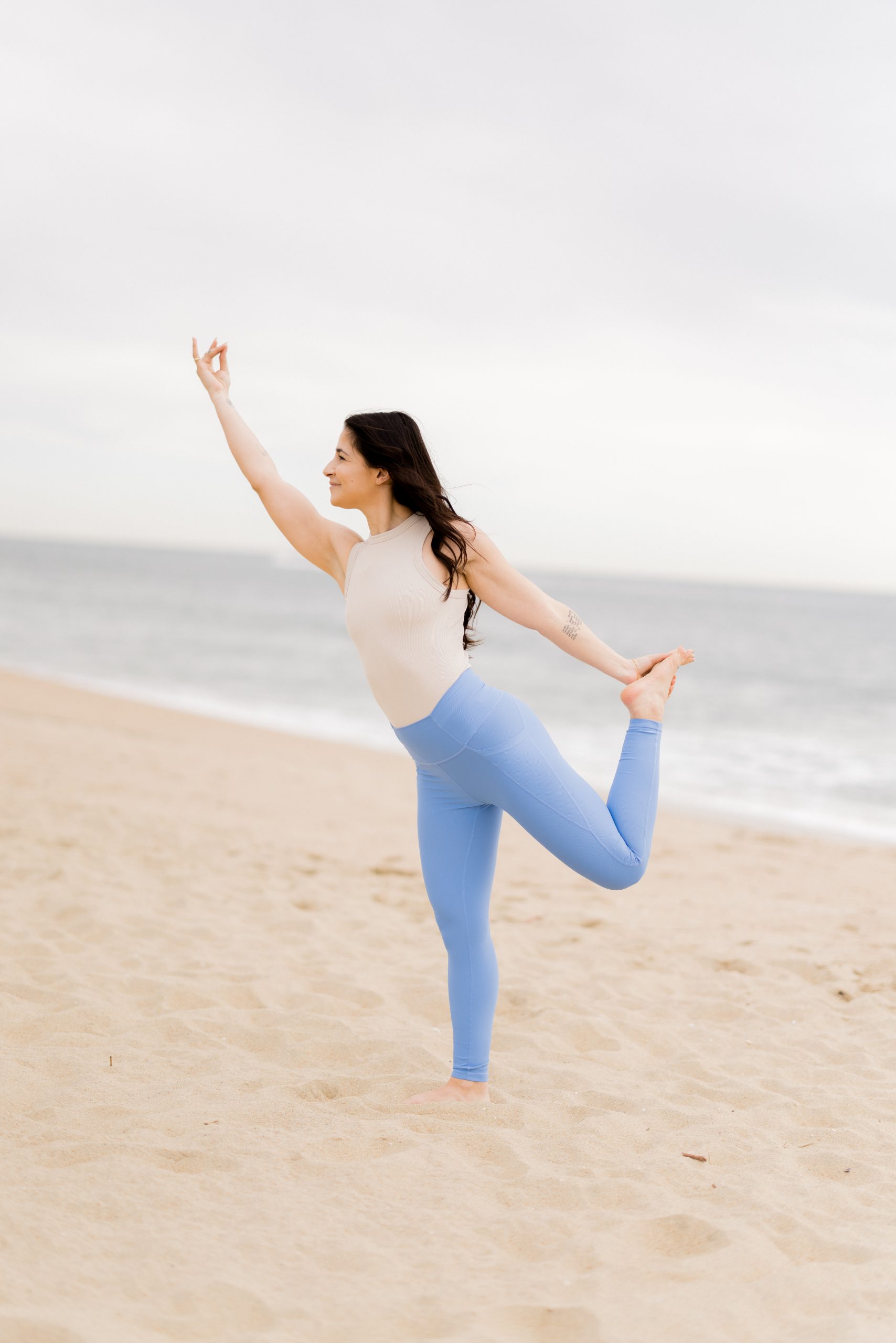 Yogi in light blue yoga pants and a beige top practicing Alia in Natarajasana on the beach