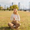 Kate Wall sitting in a meditative pose on the grass in an open park with the NYC city skyline behind her