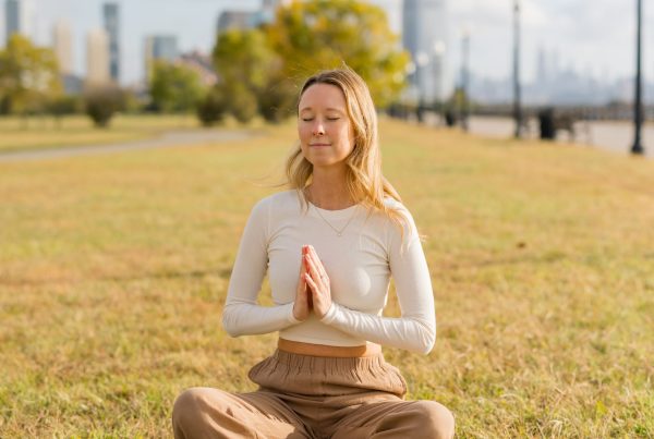 Kate Wall sitting in a meditative pose on the grass in an open park with the NYC city skyline behind her