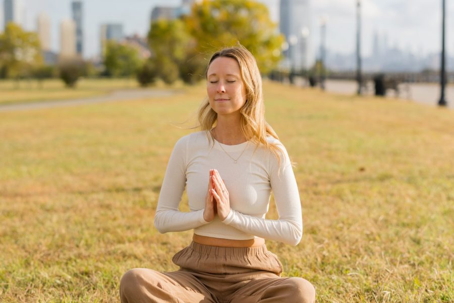 Kate Wall sitting in a meditative pose on the grass in an open park with the NYC city skyline behind her