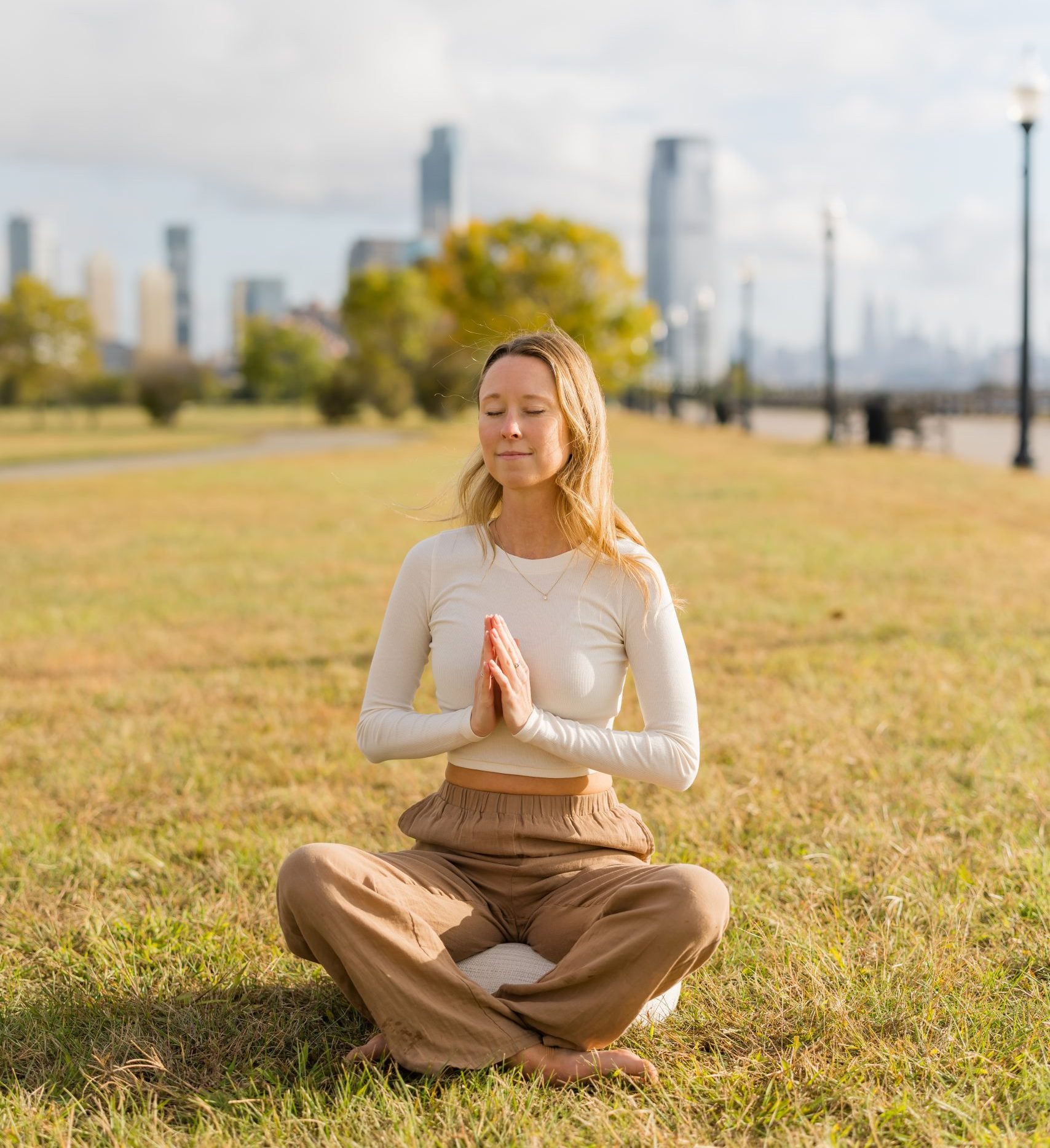 Kate Wall sitting in a meditative pose on the grass in an open park with the NYC city skyline behind her