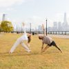 Kate and Patrick of YogaRenew doing triangle pose facing each other outside on the grass in Hoboken, NJ with the NYC skyline in the back