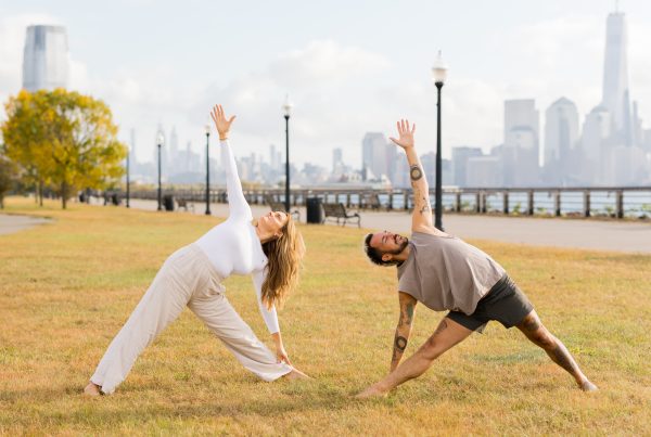 Kate and Patrick of YogaRenew doing triangle pose facing each other outside on the grass in Hoboken, NJ with the NYC skyline in the back