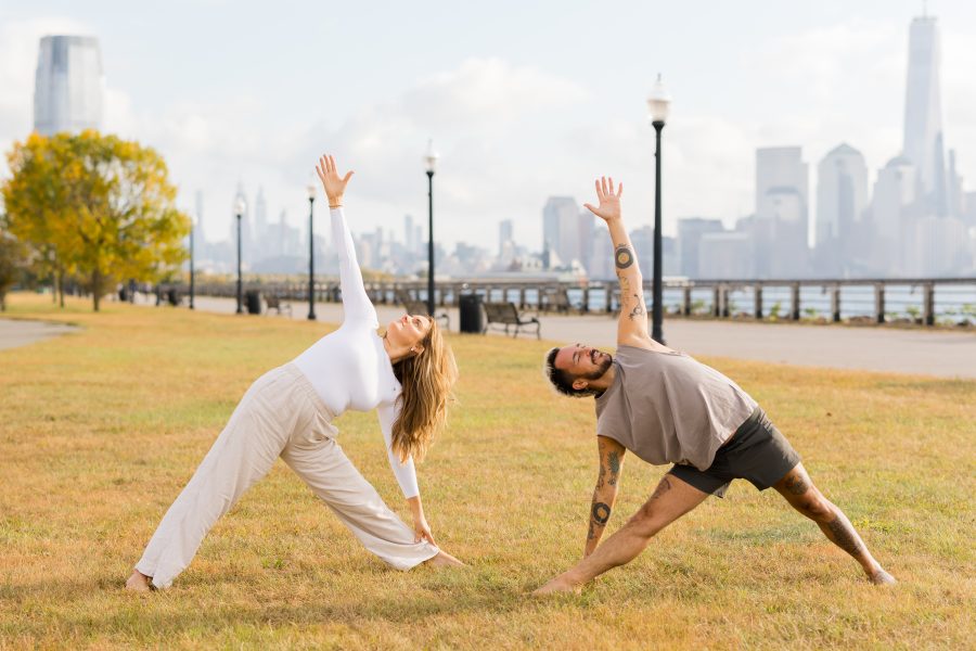 Kate and Patrick of YogaRenew doing triangle pose facing each other outside on the grass in Hoboken, NJ with the NYC skyline in the back