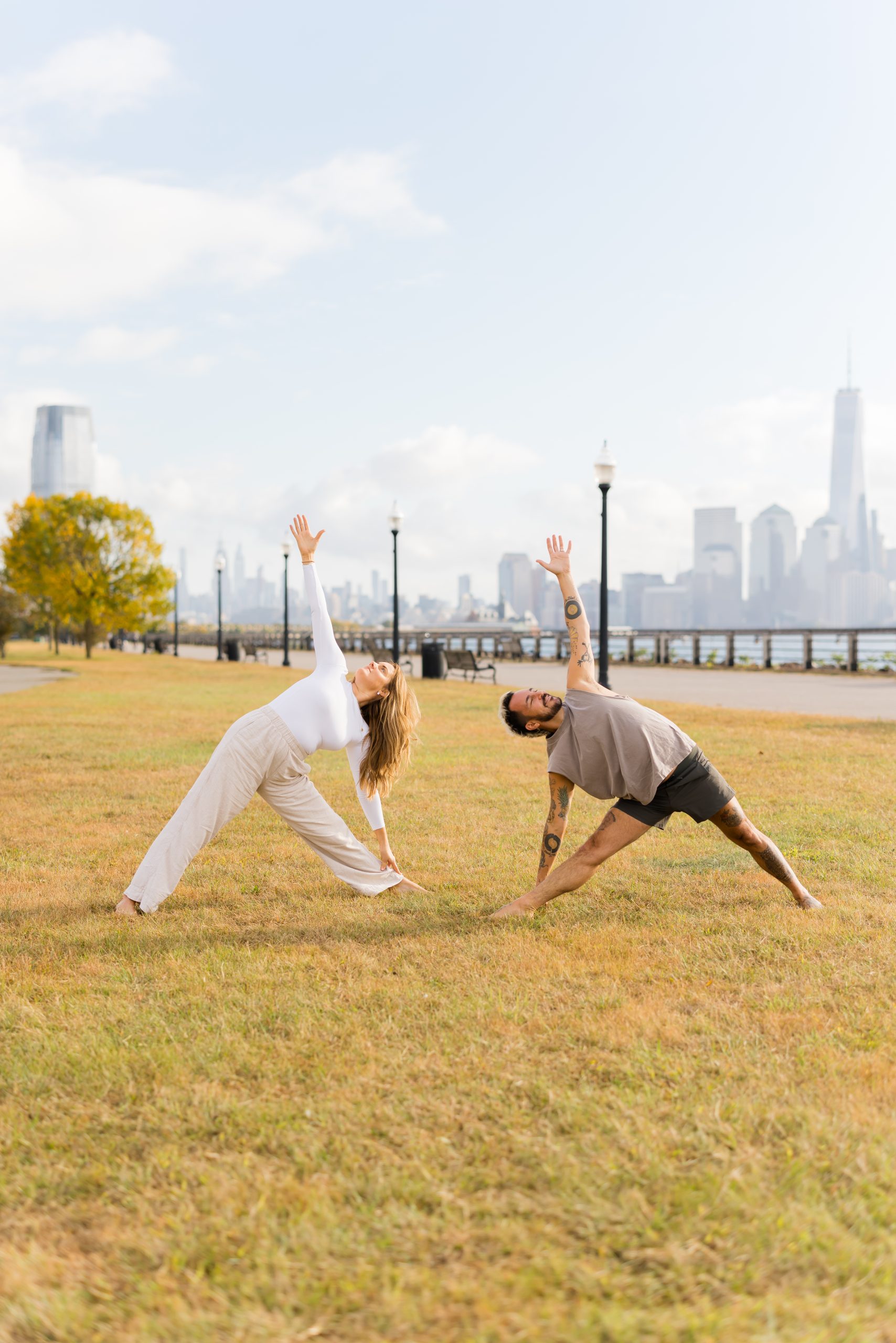 Kate and Patrick of YogaRenew doing triangle pose facing each other outside on the grass in Hoboken, NJ with the NYC skyline in the back