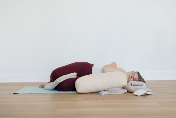 yogi resting in a restorative yoga posture with a blanket between the legs, lying on the side of the body, hugging a bolster with a blanket under the head