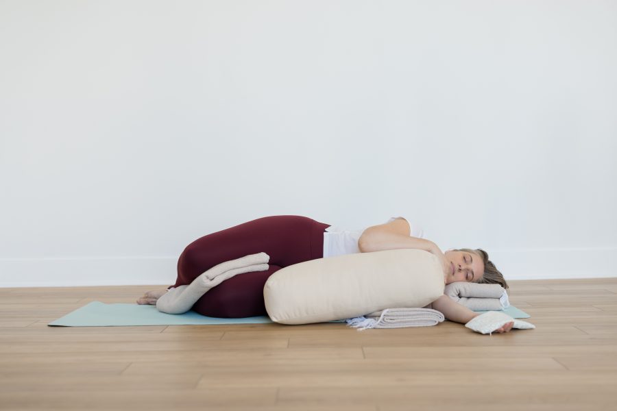 yogi resting in a restorative yoga posture with a blanket between the legs, lying on the side of the body, hugging a bolster with a blanket under the head